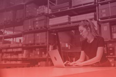 A woman in a black t-shirt working on a laptop while standing at a table in a warehouse. There are tall shelves stacked with large boxes and containers in an industrial setting. The image has a red gradient overlay, adding a modern and vibrant effect.